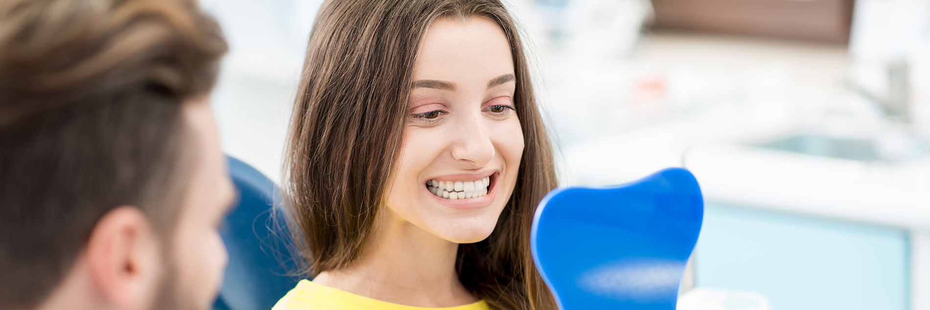 A person is seated in a dental chair, receiving care from a dental professional who stands behind them.